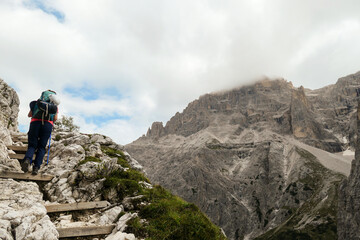 Woman with big backpack and sticks, walking up a wooden stairs in high Italian Dolomites. There are many sharp peaks in front of her. She is going up. There are a few trees around. Sunny day. Outdoor