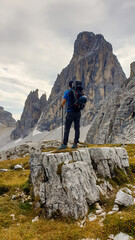 Man with big backpack and sticks, hiking in high Italian Dolomites. There are many sharp peaks in front of him. He is standing on a big boulder. Lots of lose stones and landslides. Sunny day. Outdoor