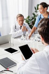 Digital tablet with blank screen in hands of doctor near multiethnic colleagues and devices on blurred background