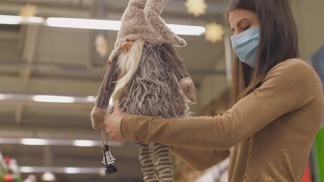 Shopaholic in face mask holds brown gnome toy choosing present for friend in local department store. Concept hobby leisure