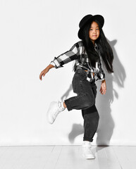 Naklejka premium Portrait of an Asian girl with black hair who dances and has fun on a white background. The child wears a black hat, plaid shirt, jeans and looks into the camera. Children's fashion and style.