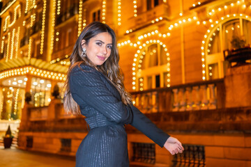 Fototapeta premium Christmas shooting, Caucasian brunette girl smiling in the city at night next to the Christmas lights. Elegant black christmas or new year's eve dress with black boots