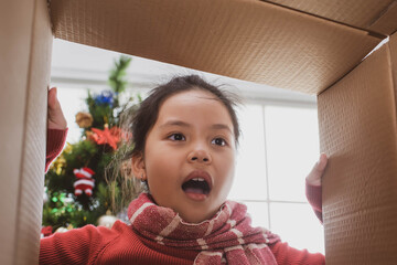 merry christmas and happy holidays. cheerful cute child girl opening a christmas present. little kid having fun near christmas tree indoors. view from inside of the box.
