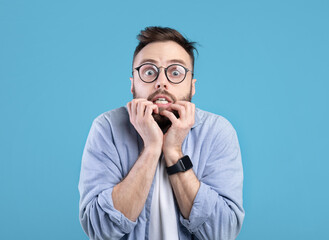 Scared millennial guy biting nails in panic over blue studio background