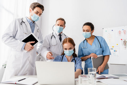 Multiethnic Doctors And Nurses In Medical Masks Using Laptop While Working Near Papers On Blurred Foreground