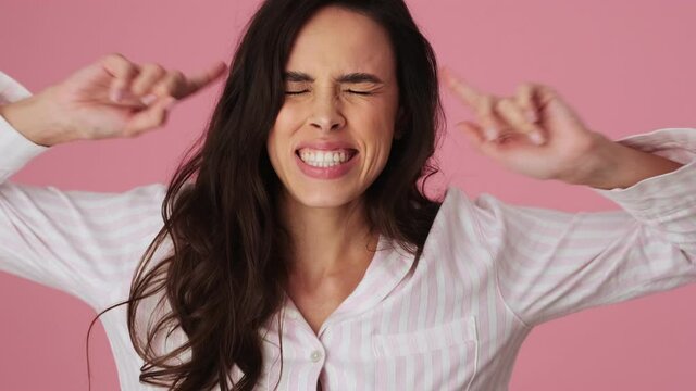 An emotional young woman is hiding her ears with her fingers and screaming standing isolated over a pink background in the studio