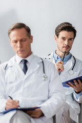 Thoughtful doctor with pen and clipboard looking at camera during meeting with blurred colleague on foreground in hospital