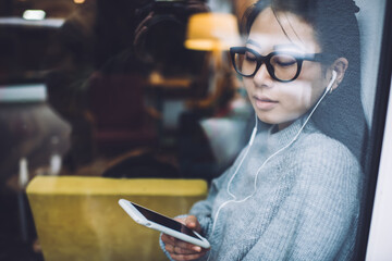 Relaxed Asian woman with phone and earphones on windowsill in cafe