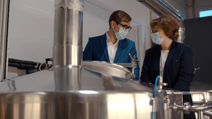 Young male manager in suit and safety mask showing beer fermentation tank to female inspector