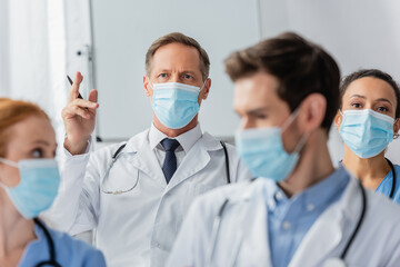 Doctor with raised hand near african american nurse during meeting with blurred colleagues on foreground in hospital