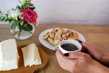 Obraz na płótnie Canvas Cup of coffee in female hands, close-up. Homemade baked goods during quarantine. Homemade feta cheese on a wooden tabletop. Concept on the background of a white brick wall and a bouquet of roses.