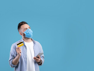 Fototapeta na wymiar Millennial man in face mask holding smartphone and credit card, looking at empty space on blue studio background