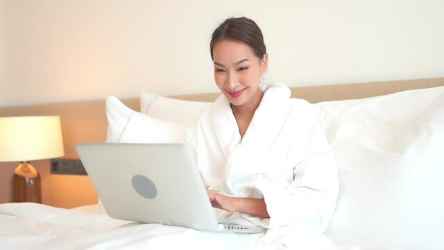 A pretty young woman works from anywhere on her laptop in a comfortable hotel bed surrounded by pillows.