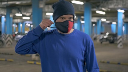 Caucasian man in a hat stands in an underground Parking lot and puts on a red and black mask against the virus