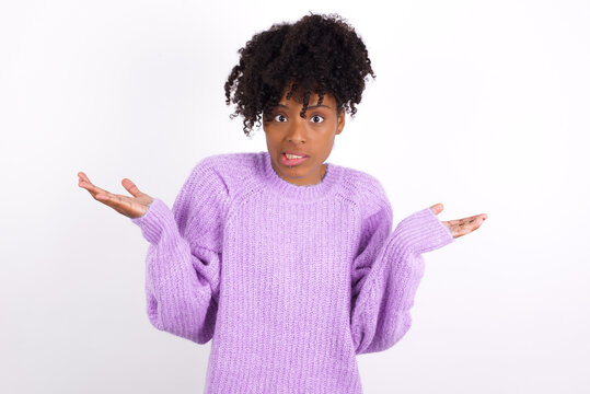Clueless African American woman wearing purple knitted sweater against white wall shrugs shoulders with hesitation, faces doubtful situation, spreads palms, Hard decision