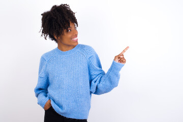 Smiling oung beautiful African American woman wearing blue knitted sweater against white wall indicating finger empty space showing best low prices