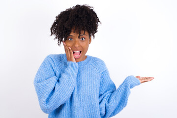 Crazy oung beautiful African American woman wearing blue knitted sweater against white wall advising discount prices hold open palm new product