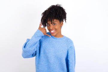 Young beautiful African American woman wearing blue knitted sweater against white wall smiling doing phone gesture with hand and fingers like talking on the telephone. Communicating concepts.