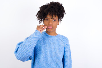 Young beautiful African American woman wearing blue knitted sweater against white wall mouth and lips shut as zip with fingers. Secret and silent, taboo talking.