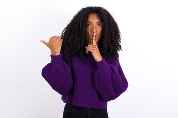 Young beautiful African American woman wearing knitted sweater against white wall,  asking to be quiet with finger on lips pointing with hand to the side. Silence and secret concept.