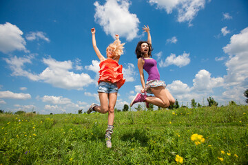 Two happy beautiful girls blonde and brunette twenty years old in colored dresses are jumping on a green field 