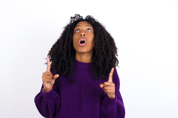 Young beautiful African American woman wearing knitted sweater against white wall amazed and surprised looking up and pointing with fingers and raised arms.