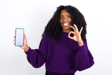 Young beautiful African American woman wearing knitted sweater against white wall holding in hands...