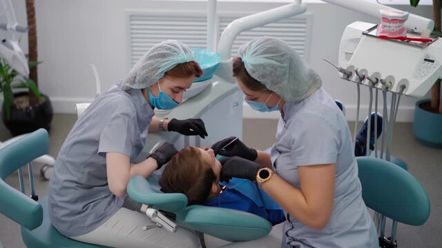 Two Woman Doctors in Medical Cap in a Dental Clinic, Serve the Patient Little Boy with Damaged Baby Teeth. Work of a Dentist with Children. Healthcare and Medicine Concept.