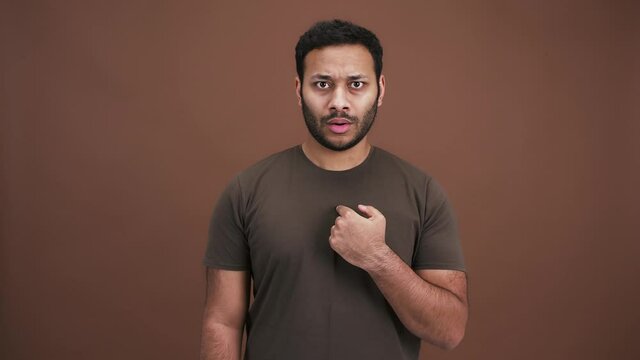 Surprised indian man looking around, pointing at himself with question and shaking head no, brown studio background