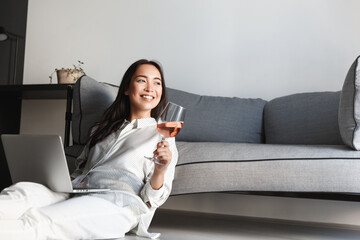 Beautiful asian woman drinking wine and using laptop, sitting on floor and resting at home