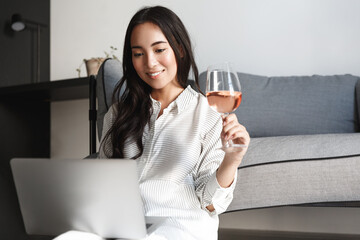 Elegant asian woman sitting at home with glass of wine and laptop, browsing online and resting