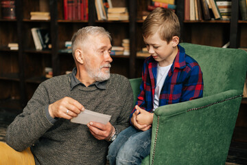 Portrait of happy bearded grandpa talking with cute grandson, having fun looking at an old photo album, enjoying memories watching family photo album at home in cozy room background on bookshelves