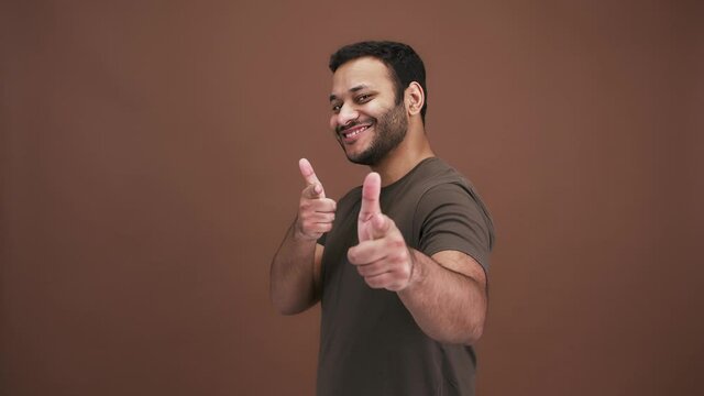 Playful indian man flirting, pointing to camera with both hands and winking, brown studio background, side view