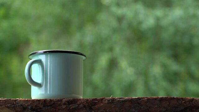 Closeup view 4k video of blue enamel mug full hot steaming drink standing on old tree log outdoors. Selective focus on cup isolated on blurry natural green foliage bokeh background.