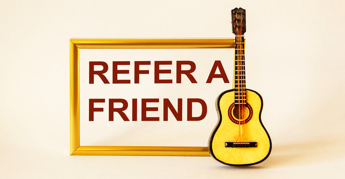 The text Refer a friend is written on a photo frame, next to a guitar on a white background. Referral marketing concept. - Powered by Adobe