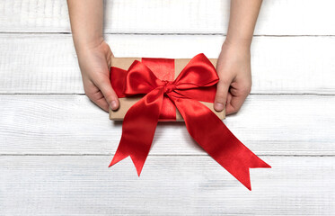 Female hands are holding a gift or real box decorated with a red ribbon with a bow on a light...