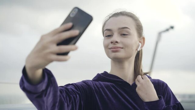 Close-up of confident beautiful sportswoman taking selfie outdoors on cloudy day. Portrait of charming young Caucasian woman taking photo after morning training outdoors.