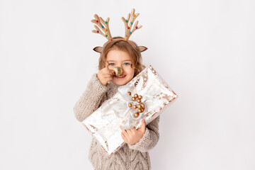 little girl with deer antlers and Christmas ball on her nose on a white background with a gift in her hands, space for text, new year and Christmas concept