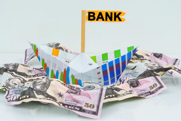 A paper boat from a business graph floats on a sea of money. The text is written on the flag - BANK