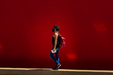 Jovem homem, caminhando na rua, usando máscara de proteção, com mochila vermelha nas costas e muro pintado de vermelho atrás.