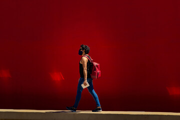Jovem homem, caminhando na rua, usando máscara de proteção, com mochila vermelha nas costas e...