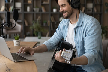 Happy millennial man musician hold play guitar watching online video or tutorial on laptop. Smiling young Caucasian male artist or singer record new music single use computer at home studio.