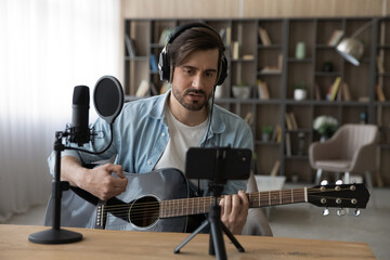Millennial Caucasian male music tutor coach hold guitar record video tutorial on smartphone at home studio. Young man artist or singer play musical instrument, shoot new song or single on cellphone.