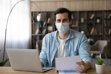 Fototapeta na wymiar Portrait of smiling young Caucasian male employee in facial mask sit at desk in office work distant on laptop. Happy man worker in medical facemask use computer busy with documents during covid-19.