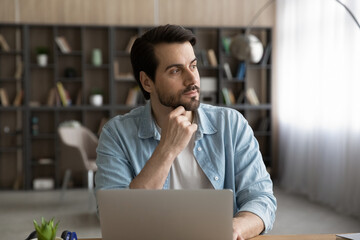 Pensive young Caucasian woman work on laptop in home office look in window distance thinking...