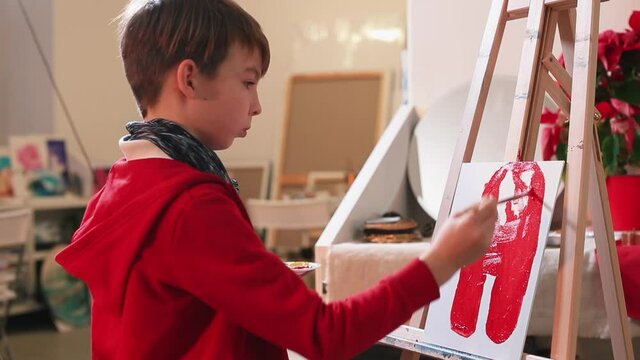The student paints a picture with red paint. He holds a brush with his hand, paints the canvas on a malbert with red paint. Getting art education.