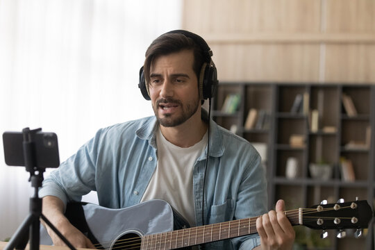 Millennial 20s Caucasian male artist or blogger in headphones play guitar record music video on smartphone at home. Young man singer or composer use musical instrument sing, compose new song on cell.