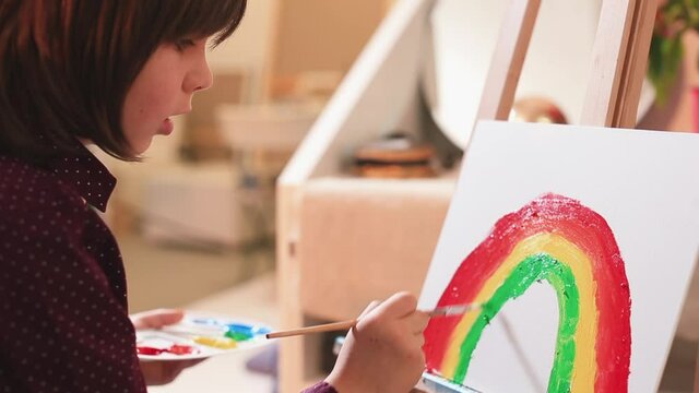A schoolboy paints a rainbow on canvas in self-isolation. He dabs green paint with a brush. The focus is on the process. Creating a picture of a rainbow trend.