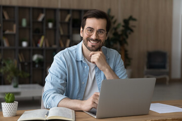 Portrait of smiling millennial Caucasian male employee in glasses sit at desk at home office...