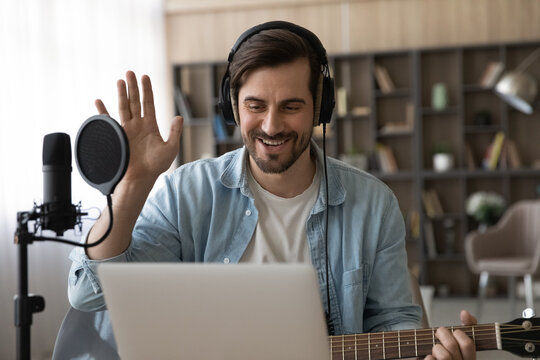 Happy millennial male artist in headphones and guitar have online video music lesson on computer. Smiling young man singer tutor or coach talk greet on webcam digital virtual training at home studio.
